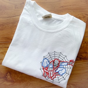 May include: White t-shirt with an embroidered image of Spiderman in a web. The embroidery is in red, blue, and black thread.