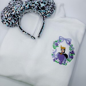 May include: White sweatshirt with an embroidered Evil Queen design in a floral wreath. A sequined black and iridescent headband with large ears rests on the sweatshirt. The design features purple, green, and gold accents.