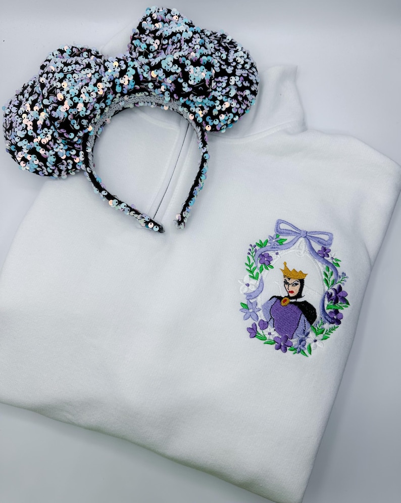 May include: White sweatshirt with an embroidered Evil Queen design in a floral wreath. A sequined black and iridescent headband with large ears rests on the sweatshirt. The design features purple, green, and gold accents.