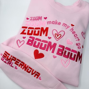 May include: A light pink sweatshirt with embroidered text and hearts. The text includes "ZOOM" and "BOOM" in shades of red and pink, along with "make my heart go" and "SUPERNOVA GIRL". The design features multiple heart shapes.