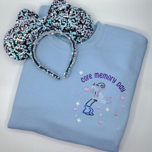 May include: A light blue sweatshirt with the text "core memory day" embroidered on the front. The sweatshirt is paired with a black and iridescent sequined headband with large bow ears.