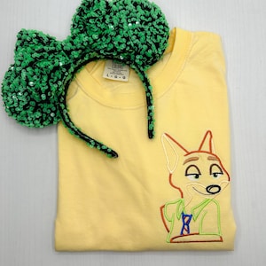 May include: A yellow t-shirt with an embroidered image of Nick Wilde from the movie Zootopia. The shirt is paired with a green sequin headband with black elastic.