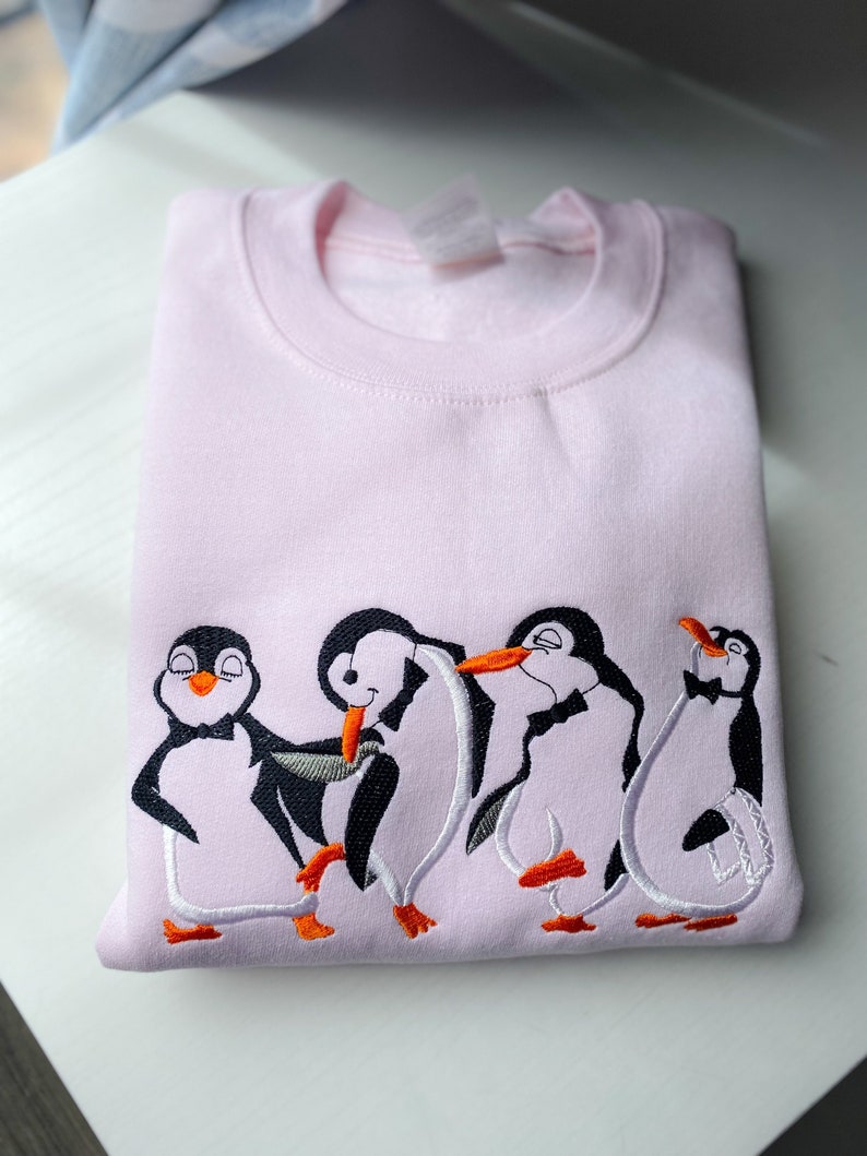 May include: A pink sweatshirt with an embroidered design of four penguins wearing bowties. The penguins are in a line, with the first two penguins facing each other and the last two penguins facing forward. The penguins are embroidered in white, black, and orange.