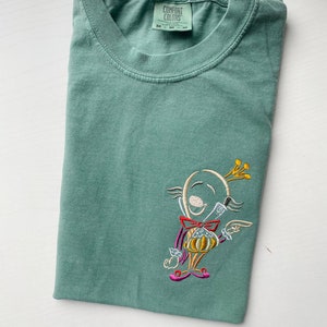 May include: A teal green t-shirt with an embroidered cartoon character wearing a crown and holding a pumpkin. The t-shirt has a tag that reads "COMFORT COLORS" with the size "M" printed on it.