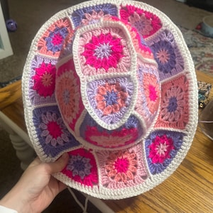 May include: A vibrant crochet hat with a patchwork design in pink, purple, and white. The hat has a wide brim and a drawstring. The crochet work is detailed, showcasing a floral pattern.