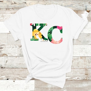 May include: White t-shirt with a floral print design of the letters "KC" with the words "Born and Loyal" underneath.