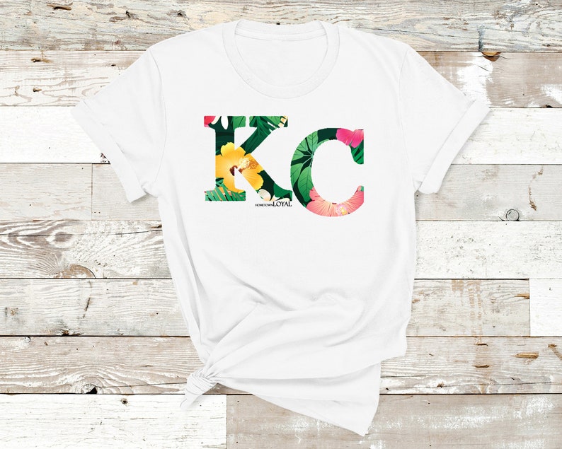 May include: White t-shirt with a floral print design of the letters "KC" with the words "Born and Loyal" underneath.