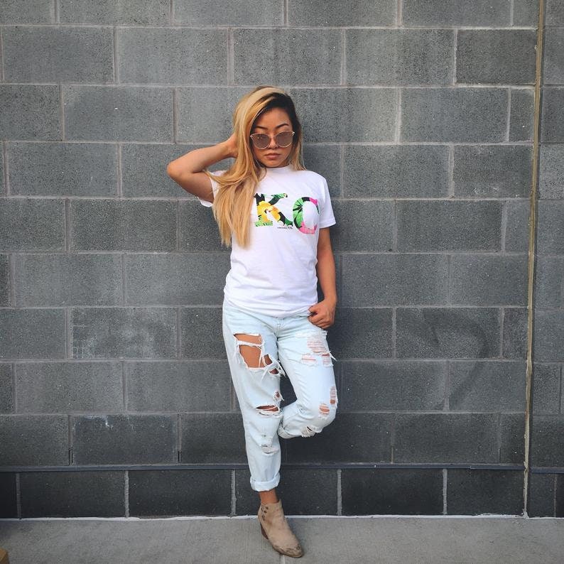 May include: A woman wearing a white t-shirt with a floral design that spells out "KC". She is wearing light blue ripped jeans and brown ankle boots. She is standing in front of a gray brick wall.