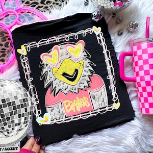 Pode incluir: Camiseta preta com uma &aacute;guia de desenho animado usando &oacute;culos de sol em forma de cora&ccedil;&atilde;o e a palavra "Eagles". Um copo xadrez rosa e branco com uma al&ccedil;a e canudo rosa est&aacute; ao lado da camiseta. Uma bola de discoteca e acess&oacute;rios cor-de-rosa tamb&eacute;m est&atilde;o na imagem.