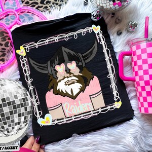 Pode incluir: Camiseta preta com um gr&aacute;fico de um viking com &oacute;culos de sol em forma de cora&ccedil;&atilde;o e a palavra "Raiders". Um copo xadrez rosa e branco com uma al&ccedil;a e canudo rosa tamb&eacute;m &eacute; vis&iacute;vel. Uma bola de discoteca tamb&eacute;m est&aacute; presente.