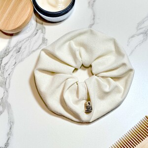May include: A white fabric scrunchie with a small tag that says "Naturally Handmade". The scrunchie is on a white marble surface with a wooden comb and a jar of cream in the background.