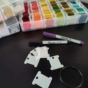May include: Two clear plastic storage containers filled with colorful embroidery floss wound on white plastic bobbins. Included are a purple Sharpie marker, a black Sharpie marker, several empty white plastic bobbins, and a black metal binder ring.