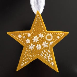 May include: A yellow felt star with white embroidery of various floral and geometric designs. The star is hanging from a white ribbon.