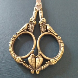 May include: Ornate gold-toned scissors with a floral design and two birds on each side. The scissors have a silver blade.
