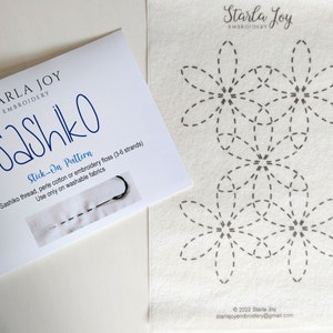 May include: A white card with the text "STARLA JOΥ EMBROIDERY Sashiko Stick-On Pattern" and a small image of a needle and thread. The card is next to a white fabric with three black sashiko stitch patterns of flowers.