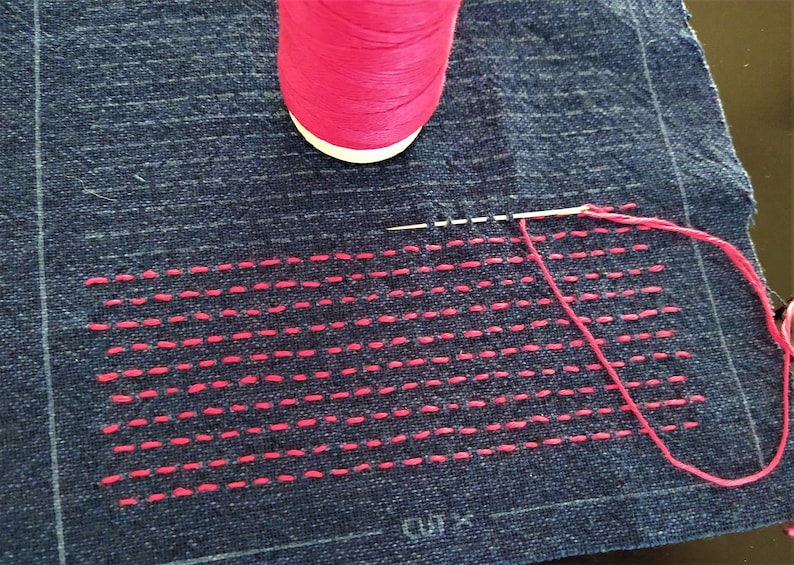 Op de afbeelding: Een close-up van een stuk blauwe stof met een roze draad gestikt in een recht lijnenpatroon. Een spoel roze draad bevindt zich in de linkerbovenhoek van de afbeelding.