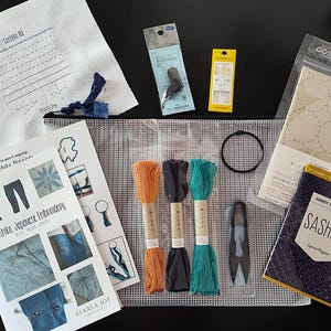 K&ouml;nnte beinhalten: Ein Sashiko-Stickset mit verschiedenen Materialien. Das Set enth&auml;lt Stickgarn in Orange, Schwarz und T&uuml;rkis, Nadeln, einen Stickrahmen, eine Anleitung und Stoff mit aufgedruckten Mustern. Das Set enth&auml;lt auch ein B&uuml;chlein mit dem Titel "Sashiko" von Sylvie Pippen.