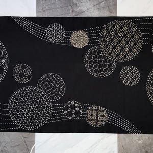 May include: Black rectangular table runner with a repeating pattern of embroidered circles in various sizes and designs. The circles are white and beige, and the background is black. The runner is approximately 12 inches wide and 36 inches long.