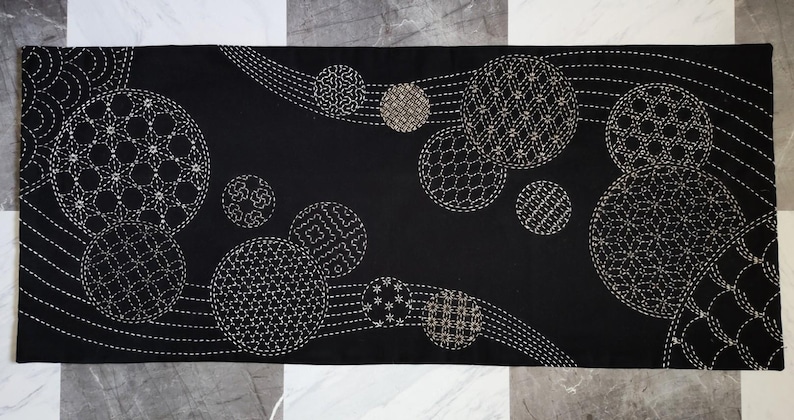 Puede incluir: Camino de mesa rectangular negro con un patr&oacute;n repetido de c&iacute;rculos bordados de varios tama&ntilde;os y dise&ntilde;os. Los c&iacute;rculos son blancos y beige, sobre un fondo negro. El camino de mesa mide aproximadamente 30 cm de ancho y 91 cm de largo.