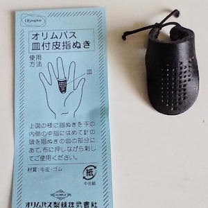 K&ouml;nnte beinhalten: Ein schwarzer Lederfingerhut mit Gittermuster und Schnur, neben einer hellblauen Karte mit japanischem Text und einer Handzeichnung. Die Karte ist mit "Olympus" beschriftet.