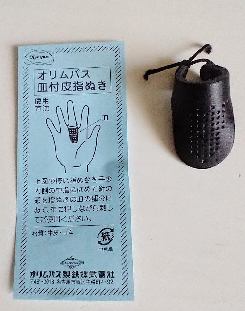 K&ouml;nnte beinhalten: Ein schwarzer Lederfingerhut mit Gittermuster und Schnur, neben einer hellblauen Karte mit japanischem Text und einer Handzeichnung. Die Karte ist mit "Olympus" beschriftet.