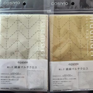 K&ouml;nnte beinhalten: Zwei verpackte Stoffquadrate mit einem gesteppten Zickzackmuster. Der Stoff ist beige und olivgr&uuml;n, mit dem Text "hidamari" und japanischem Text auf der Verpackung. Jedes Quadrat misst ca. 32 x 32 cm.
