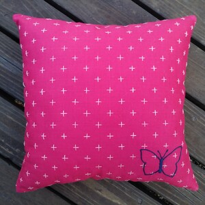 K&ouml;nnte beinhalten: Ein quadratisches Zierkissen mit leuchtend pinkfarbenem Hintergrund und einem sich wiederholenden Muster aus kleinen wei&szlig;en Kreuzen. Ein blauer Schmetterling ist auf der unteren rechten Ecke gestickt. Das Kissen ist wahrscheinlich aus Baumwolle oder einem &auml;hnlichen Stoff.