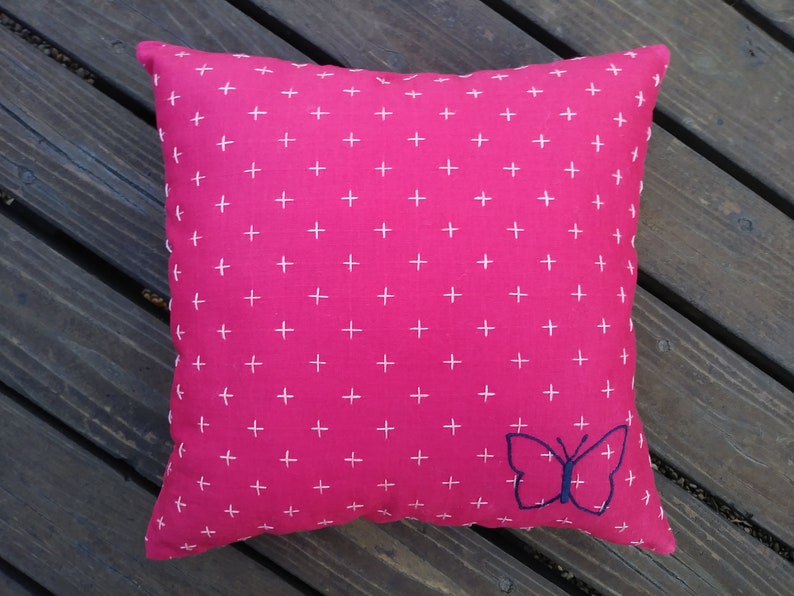 K&ouml;nnte beinhalten: Ein quadratisches Zierkissen mit leuchtend pinkfarbenem Hintergrund und einem sich wiederholenden Muster aus kleinen wei&szlig;en Kreuzen. Ein blauer Schmetterling ist auf der unteren rechten Ecke gestickt. Das Kissen ist wahrscheinlich aus Baumwolle oder einem &auml;hnlichen Stoff.