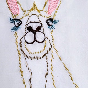 May include: Embroidered llama with pink ears, brown nose, and blue eyelashes. The llama is stitched on a white fabric background.