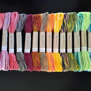 May include: A collection of sixteen skeins of variegated embroidery thread arranged in a row. Each skein is wrapped in a tan paper band labeled with the text Sashiko and Japanese characters. The threads feature a spectrum of colors including pink, red, grey, purple, orange, yellow, green, and blue.