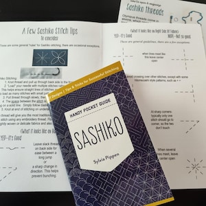 K&ouml;nnte beinhalten: Ein Bild von Anleitungen zum Sashiko-Sticken. Im Mittelpunkt steht ein "Sashiko"-Taschenf&uuml;hrer mit einem geometrischen Muster in Marineblau. Mehrere bedruckte Bl&auml;tter mit Stichdiagrammen und Tipps sind ebenfalls zu sehen.