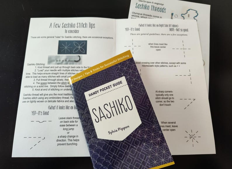 K&ouml;nnte beinhalten: Ein Bild von Anleitungen zum Sashiko-Sticken. Im Mittelpunkt steht ein "Sashiko"-Taschenf&uuml;hrer mit einem geometrischen Muster in Marineblau. Mehrere bedruckte Bl&auml;tter mit Stichdiagrammen und Tipps sind ebenfalls zu sehen.