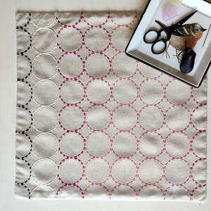 K&ouml;nnte beinhalten: Ein quadratischer, cremefarbener Stoff mit einem sich wiederholenden Kreismuster, handgestickt mit schwarzem und rosa Faden. Die Stickerei geht von Schwarz zu Rosa &uuml;ber. Eine kleine Schale mit Schere und einer Vogelillustration befindet sich in der oberen rechten Ecke.