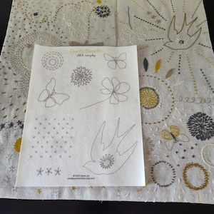 May include: A square piece of light beige fabric featuring various hand-embroidered motifs in yellow, gray, and white thread, overlaid with a white paper pattern sheet titled Create Elements Stitch everyday, displaying line drawings of butterflies, birds, and flowers.