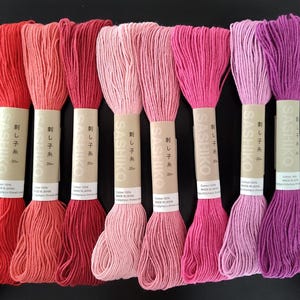 May include: A collection of eight spools of Sashiko thread in a gradient of pink and red hues. Each spool is wrapped with a paper label featuring Japanese text. The thread is made of 100% cotton and is 20 meters long.
