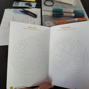 K&ouml;nnte beinhalten: Offenes Buch mit Sashiko-Designs, das Wellen- und Wirbelmuster mit gepunkteten Linien zeigt. Der Text lautet "Reduzieren oder vergr&ouml;&szlig;ern auf die gew&uuml;nschte Gr&ouml;&szlig;e." Schere, Faden und andere Bastelmaterialien sind sichtbar.