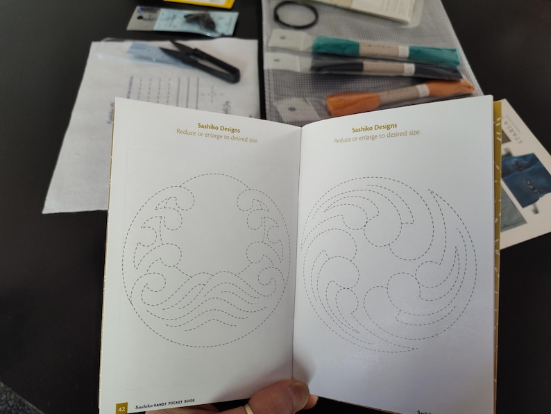 K&ouml;nnte beinhalten: Offenes Buch mit Sashiko-Designs, das Wellen- und Wirbelmuster mit gepunkteten Linien zeigt. Der Text lautet "Reduzieren oder vergr&ouml;&szlig;ern auf die gew&uuml;nschte Gr&ouml;&szlig;e." Schere, Faden und andere Bastelmaterialien sind sichtbar.