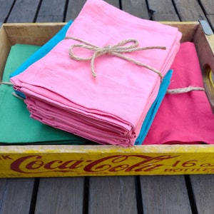 May include: A wooden Coca-Cola crate holds stacks of colorful cloth napkins. Pink, teal, and green napkins are tied with twine. The crate is yellow with red Coca-Cola branding.