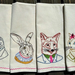 May include: Four white tea towels with embroidered images of a bird, rabbit, fox, and cat wearing suits and ties. The bird is wearing a bow tie, the rabbit has a ruffled collar, the fox has a pink tie, and the cat has a star pin.