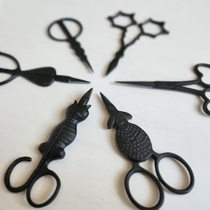 May include: A collection of six black scissors in various decorative designs. The scissors include shapes such as a heart, a cat, and a fish. The scissors are arranged in a circular pattern on a white surface.