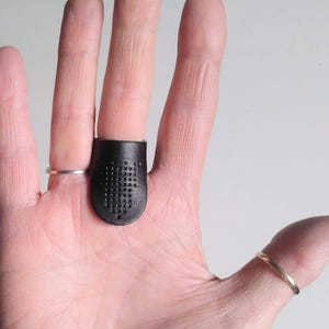 K&ouml;nnte beinhalten: Ein schwarzer Lederfingerhut mit einem Gitter aus kleinen L&ouml;chern, der an einem Finger getragen wird. Ein silberner Ring befindet sich am angrenzenden Finger. Ein goldener Ring befindet sich am Daumen. Der Fingerhut ist zum N&auml;hen bestimmt und sch&uuml;tzt den Finger vor Nadelstichen.