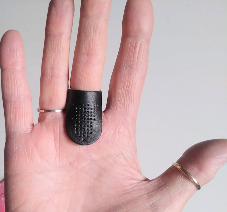 K&ouml;nnte beinhalten: Ein schwarzer Lederfingerhut mit einem Gitter aus kleinen L&ouml;chern, der an einem Finger getragen wird. Ein silberner Ring befindet sich am angrenzenden Finger. Ein goldener Ring befindet sich am Daumen. Der Fingerhut ist zum N&auml;hen bestimmt und sch&uuml;tzt den Finger vor Nadelstichen.