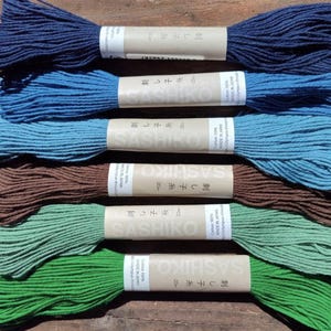 May include: Six skeins of Sashiko thread in various colors: navy blue, light blue, teal, brown, light green, and green. Each skein is wrapped with a paper label that includes Japanese text and the word "Sashiko". The threads are arranged on a wooden surface.