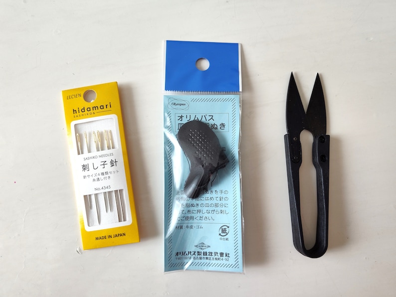 Op de afbeelding: Een set van drie gele en gouden naalden in een doorzichtige plastic verpakking met een geel label waarop "SASHIKO NEEDLES 刺し子針" en "MADE IN JAPAN" staat. Een blauwe en witte verpakking met een zwarte vingerhoed en een klein zwart schaartje.