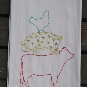 May include: A white kitchen towel with embroidered images of a chicken, pig, and cow. The chicken is green, the pig is yellow with a white outline, and the cow is red.