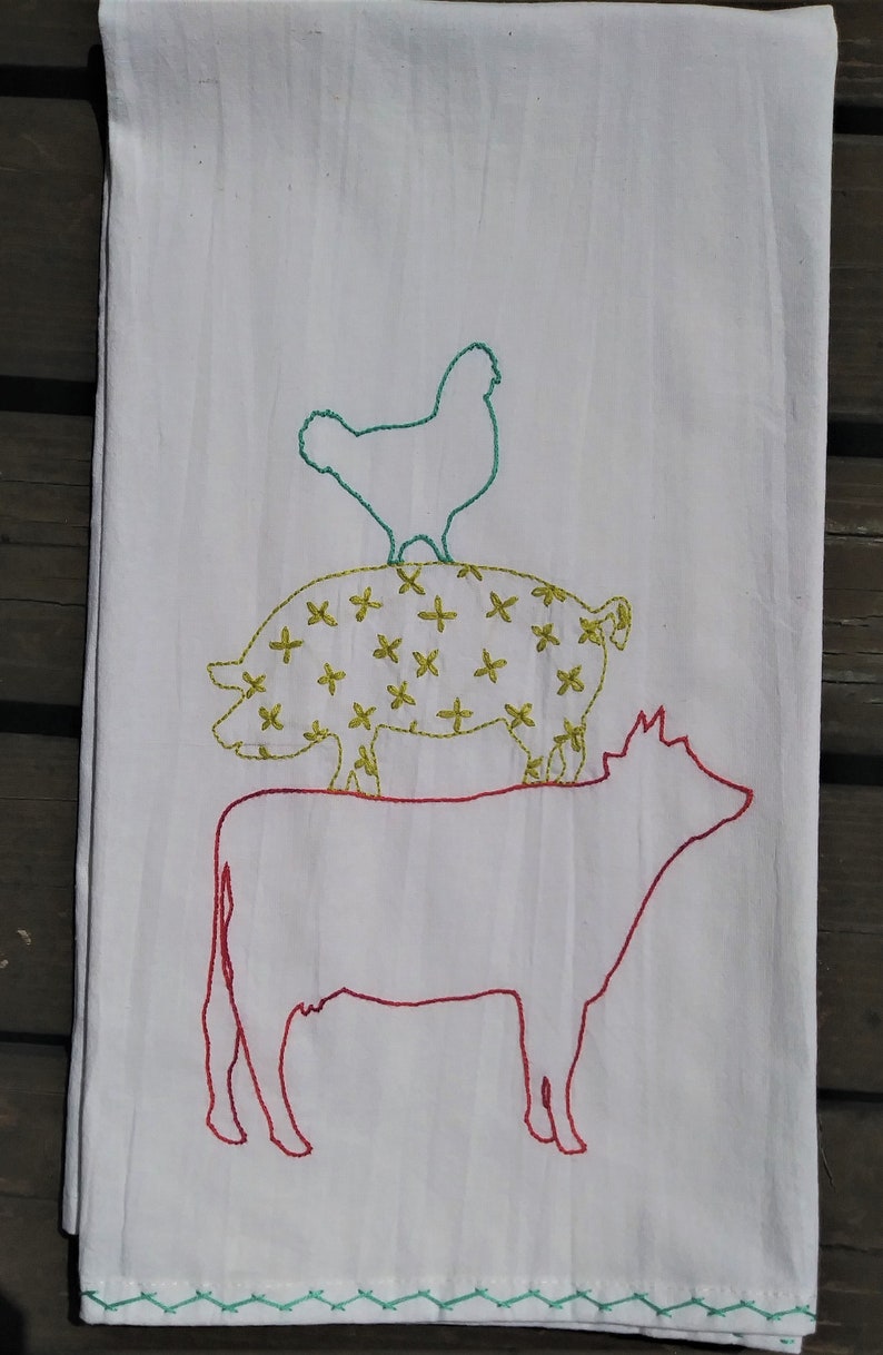 May include: A white kitchen towel with embroidered images of a chicken, pig, and cow. The chicken is green, the pig is yellow with a white outline, and the cow is red.