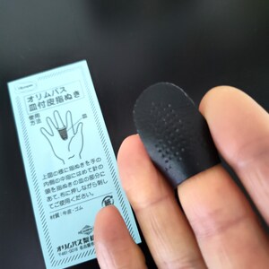 Op de afbeelding: Een zwarte vingerhoed met een getextureerd oppervlak. De vingerhoed wordt op een vinger gedragen en wordt gebruikt voor het naaien. De afbeelding bevat ook een blauwe en witte instructiekaart met Japanse tekst.