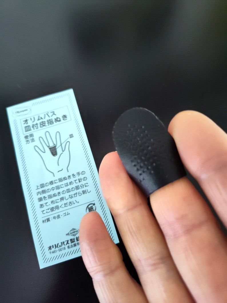 Op de afbeelding: Een zwarte vingerhoed met een getextureerd oppervlak. De vingerhoed wordt op een vinger gedragen en wordt gebruikt voor het naaien. De afbeelding bevat ook een blauwe en witte instructiekaart met Japanse tekst.