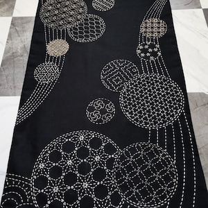 Puede incluir: Camino de mesa textil negro con patrones circulares bordados en blanco y beige. El dise&ntilde;o presenta c&iacute;rculos de varios tama&ntilde;os con intrincadas costuras geom&eacute;tricas y florales. El camino de mesa se muestra sobre un suelo a cuadros.