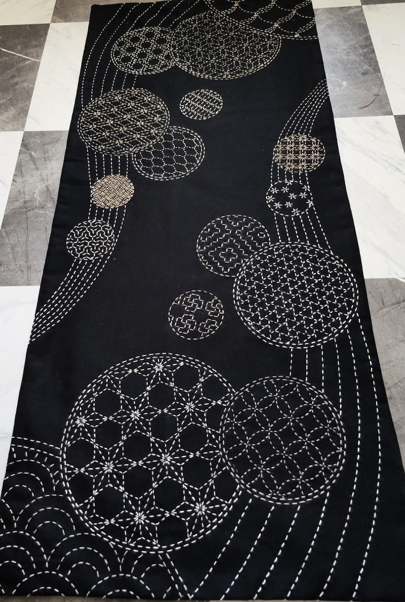 Puede incluir: Camino de mesa textil negro con patrones circulares bordados en blanco y beige. El dise&ntilde;o presenta c&iacute;rculos de varios tama&ntilde;os con intrincadas costuras geom&eacute;tricas y florales. El camino de mesa se muestra sobre un suelo a cuadros.
